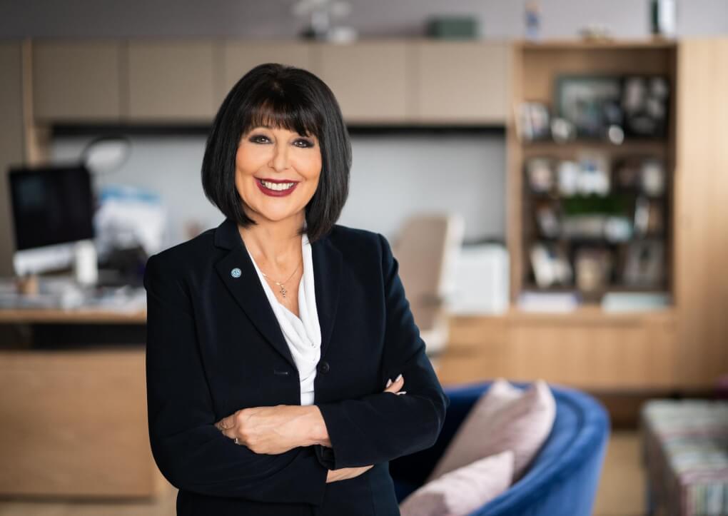 Philomena Mantella smiles, wearing a dark blue suit with Grand Valley Pin. A desk, computer, bookshelves with pictures, and armchairs are blurred out in the background.